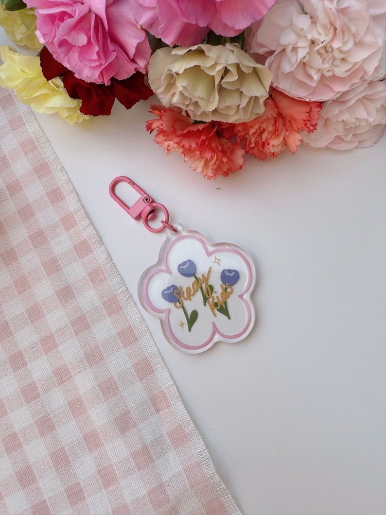 Pu&ograve; includere: Un portachiavi bianco a forma di fiore con un contorno rosa. Il portachiavi ha due fiori blu con steli verdi e una scritta dorata che dice "Sleepy Kids".