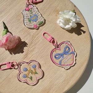 Pu&ograve; includere: Quattro portachiavi in ​​acrilico trasparente e rosa con diversi design. I portachiavi presentano un cuore con un nastro blu e il testo "Tomorrow Together", un fiore con il testo "Cherry Blossoms", una borsa a rete con il testo "Hope", e un fiore con il testo "Cherry Blossoms".