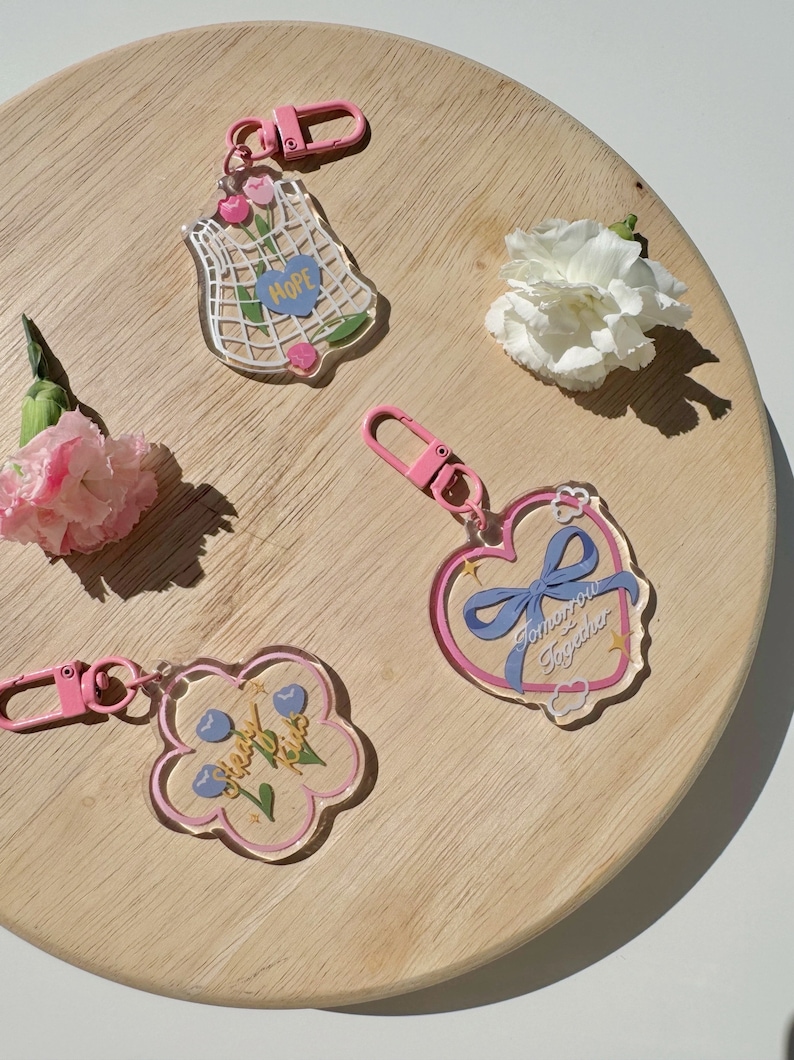 Pu&ograve; includere: Quattro portachiavi in ​​acrilico trasparente e rosa con diversi design. I portachiavi presentano un cuore con un nastro blu e il testo "Tomorrow Together", un fiore con il testo "Cherry Blossoms", una borsa a rete con il testo "Hope", e un fiore con il testo "Cherry Blossoms".