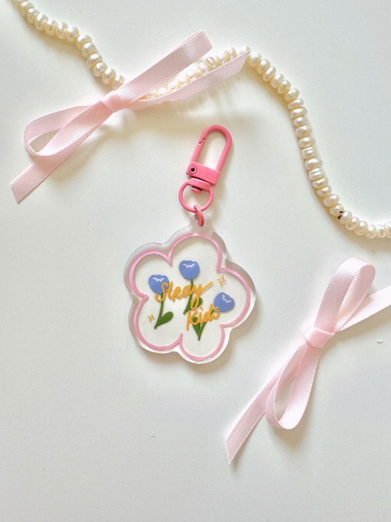Pu&ograve; includere: Un portachiavi in acrilico trasparente con un contorno rosa a forma di fiore. Il fiore ha tre fiori blu e una scritta dorata che dice "Sleepy Kids". Il portachiavi ha un gancio rosa e argentato.