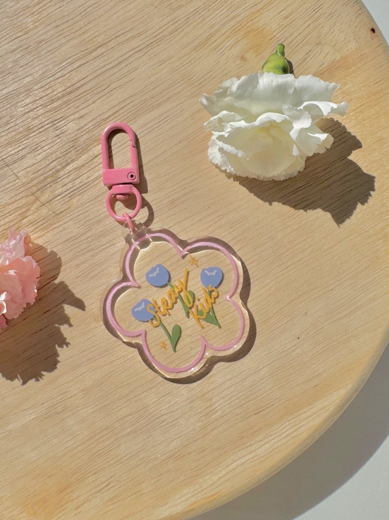 Pu&ograve; includere: Un portachiavi a forma di fiore rosa e trasparente con un moschettone in metallo rosa. Il portachiavi ha un'iscrizione gialla che recita "Steady Kids" con fiori blu e foglie verdi.