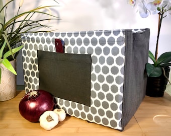 May include: A gray and white polka dot fabric storage bin with a dark gray pocket on the front. The bin is sitting on a wooden surface with a red onion and two garlic bulbs in front of it.