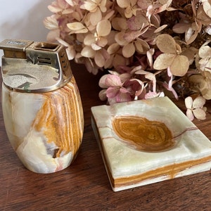 May include: A set of two matching onyx ashtrays. The larger ashtray is square with a round depression in the centre. The smaller ashtray is round and has a silver top with a hinged lid. Both ashtrays have a brown, white and gold marbled pattern.