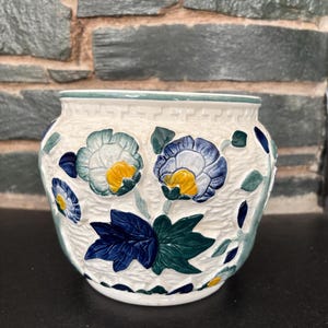 May include: A white ceramic planter with a green rim and embossed floral design. The flowers are blue, white, and yellow with green leaves.