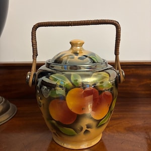 May include: A ceramic jar with a lid and a woven handle. The jar is decorated with a hand-painted design of oranges and green leaves against a black and gold background. The jar is sitting on a wooden surface.