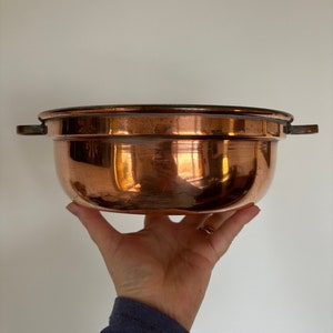 May include: A copper bowl with two handles. The bowl is shiny and has a rounded bottom.
