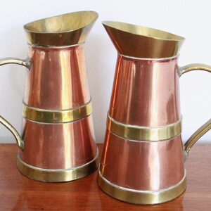 May include: Two copper jugs with brass accents. The jugs have a flared top and a rounded bottom. The handles are made of brass.