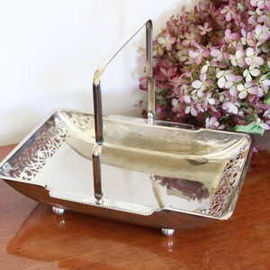 May include: Silver serving tray with a decorative cut-out design and a handle. The tray has a rectangular shape and sits on four small feet.