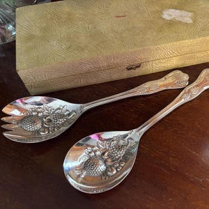 May include: Two silver serving spoons with decorative handles and fruit and leaf patterns on the spoon bowls. They are presented on a dark wooden surface, with a vintage green box visible in the background.