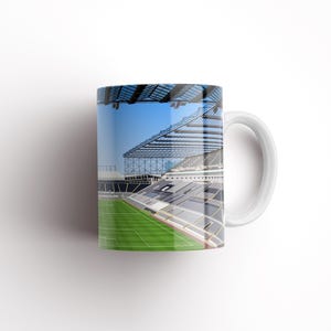 Op de afbeelding: Witte keramische mok met een kleurrijk beeld van een voetbalstadion. Het stadion heeft een groot, open dak en een groen veld.