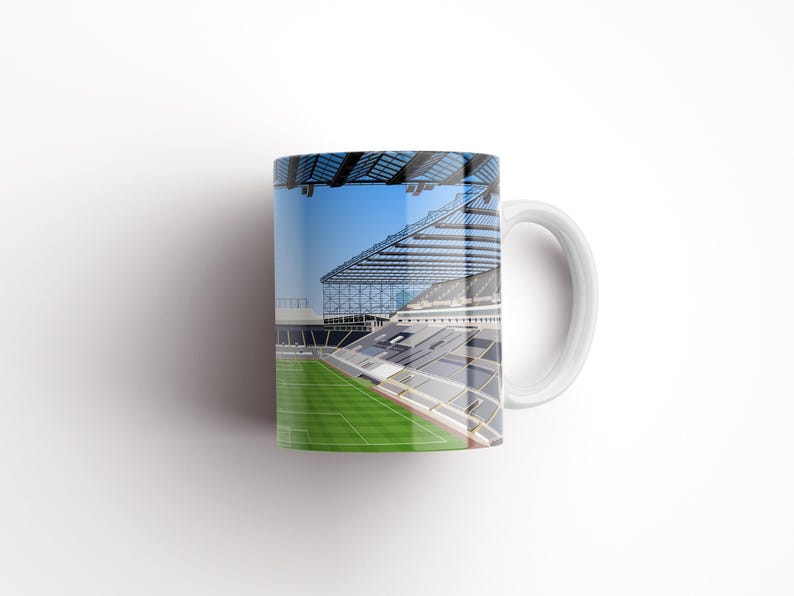 Op de afbeelding: Witte keramische mok met een kleurrijk beeld van een voetbalstadion. Het stadion heeft een groot, open dak en een groen veld.