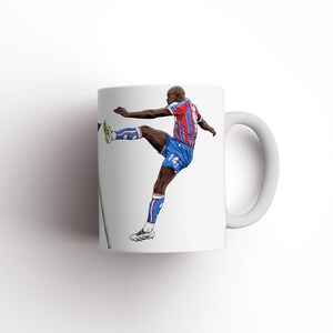 May include: White ceramic mug with a football player illustration. The player is wearing a blue, red, and white kit, mid-kick. The mug has a white handle and a glossy finish. The design is a vibrant depiction of a football action.