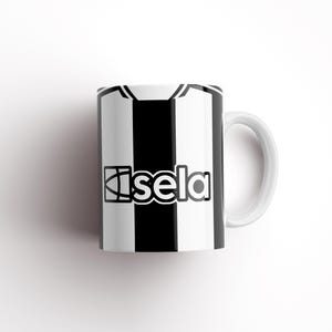 May include: White ceramic mug with a black and white striped design. The mug has a black logo that says "Isela" on the front.