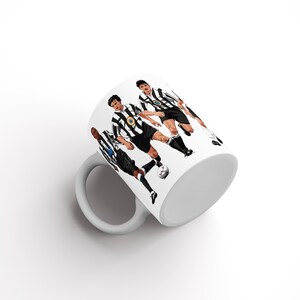 May include: White ceramic mug with a graphic of soccer players in black and white striped shirts. The players are depicted in action, kicking a football. The mug has a white handle and interior.