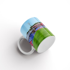 May include: White ceramic mug featuring a colourful illustration of a stadium with a green pitch and blue and red seating. The stadium is surrounded by trees and a blue sky.