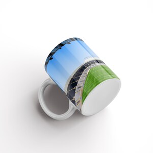Op de afbeelding: Witte keramische mok met een kleurrijk beeld van een stadion met een groen veld en een blauwe lucht.