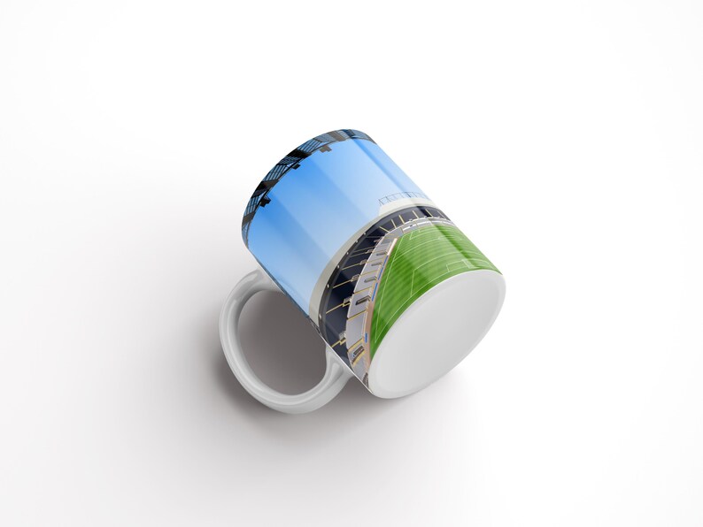 Op de afbeelding: Witte keramische mok met een kleurrijk beeld van een stadion met een groen veld en een blauwe lucht.