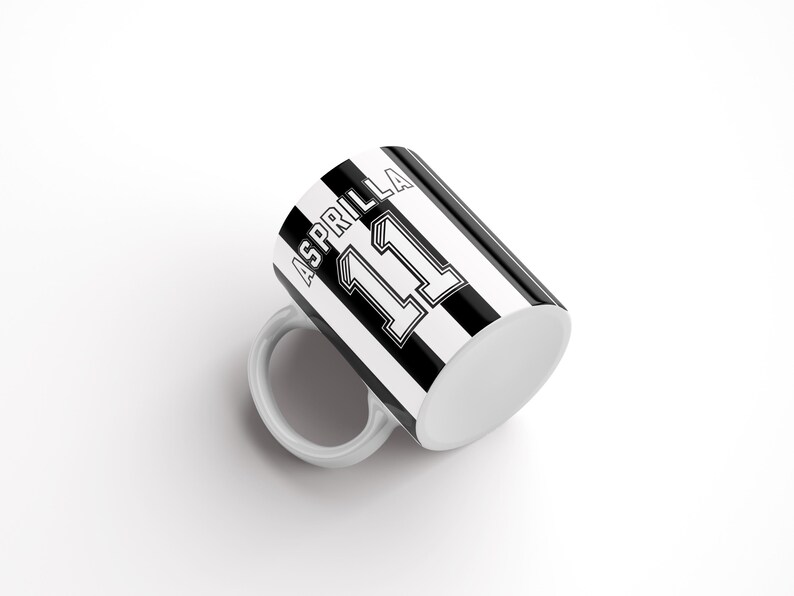 May include: A white ceramic mug with a black and white striped design. The mug has the name "Asprilla" and the number "11" printed on it.