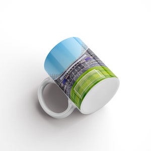 May include: White ceramic mug featuring a colourful image of a stadium with a green pitch and a goal. The mug is tilted to the side.