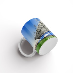 May include: White ceramic mug with a colourful image of a stadium. The image features a blue sky, a white building with a green field in the foreground. The mug has a green band around the top.