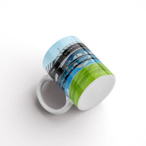 May include: White ceramic mug featuring a colourful image of a stadium and a green field. The mug is tilted to the side, revealing the bottom of the mug.