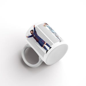 May include: White ceramic mug with a design of a soccer player in a blue and red uniform. The mug has a white handle and a white interior. The design is printed on the side of the mug.
