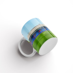 May include: White ceramic mug featuring a colourful illustration of a football stadium with a green pitch and blue stands. The mug is tilted to the side, revealing the bottom of the mug.