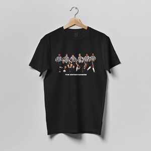 May include: Black t-shirt featuring a graphic of a soccer team in black and white striped jerseys. The text "THE ENTERTAINERS" is printed below the image. The shirt is hanging on a wooden hanger.