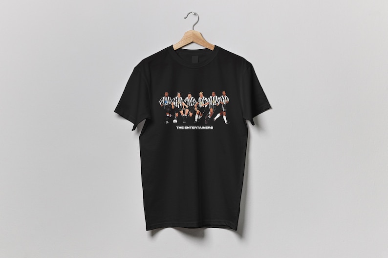 May include: Black t-shirt featuring a graphic of a soccer team in black and white striped jerseys. The text "THE ENTERTAINERS" is printed below the image. The shirt is hanging on a wooden hanger.