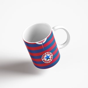 May include: White ceramic mug with a blue and red striped design. The mug features a circular logo with a star and the text "Newcastle Brown Ale".