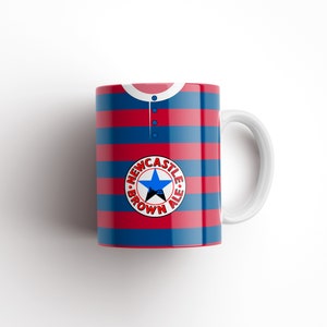 May include: A white ceramic mug with a red and blue striped design. The mug features a circular logo with the text "NEWCASTLE BROWN ALE" and a star.