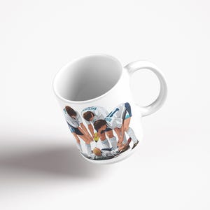 May include: White ceramic mug with a football team illustration. The players are in white and blue kits, huddled together. The mug has a curved handle and a glossy finish.