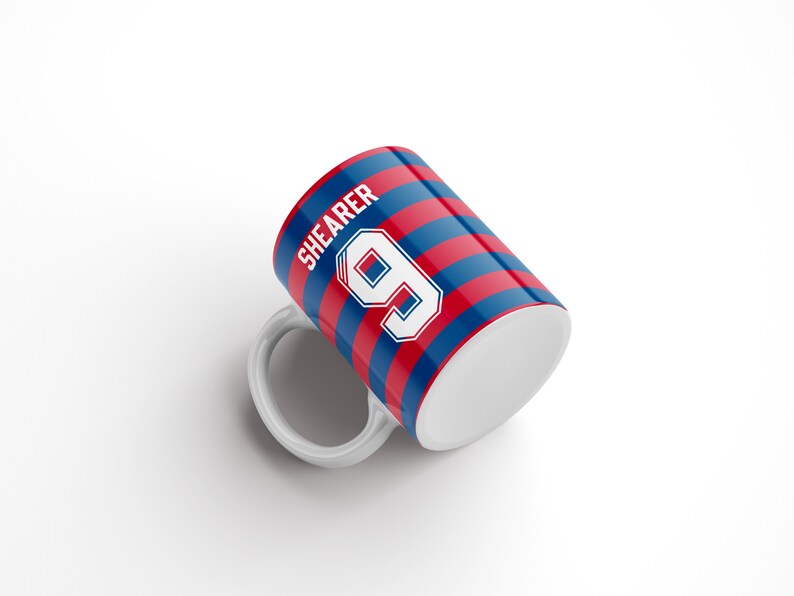 May include: White ceramic mug with a red and blue striped design. The mug features the name "SHEARER" and the number "9" in white.