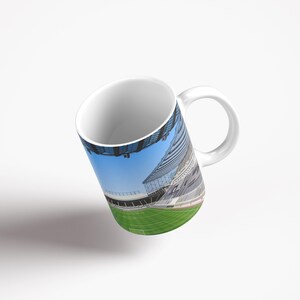 Op de afbeelding: Witte keramische mok met een full-color wrap-around afbeelding van een voetbalstadion. Het stadion is leeg en het veld is groen. De lucht is blauw.