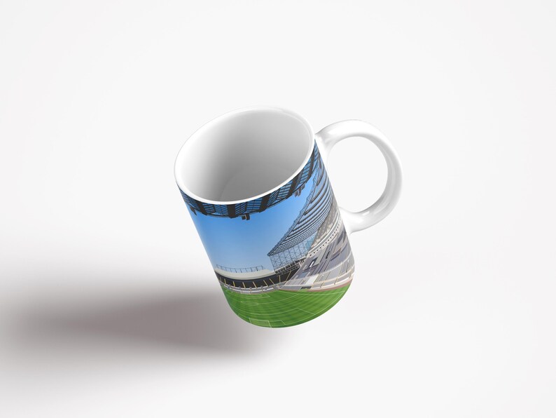 Op de afbeelding: Witte keramische mok met een full-color wrap-around afbeelding van een voetbalstadion. Het stadion is leeg en het veld is groen. De lucht is blauw.