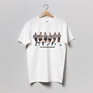 May include: White t-shirt featuring a graphic of six soccer players in black and white striped jerseys, running. The text "THE ENTERTAINERS" is printed below the players. The shirt is hanging on a wooden hanger.
