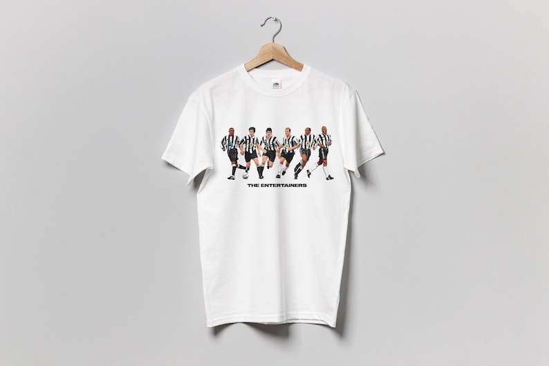 May include: White t-shirt featuring a graphic of six soccer players in black and white striped jerseys, running. The text "THE ENTERTAINERS" is printed below the players. The shirt is hanging on a wooden hanger.