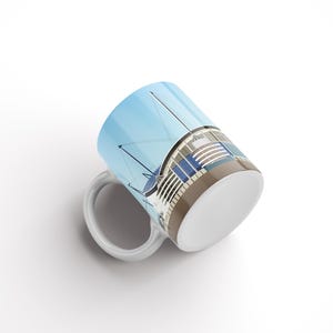 May include: White ceramic mug featuring a colourful illustration of a stadium against a blue sky.