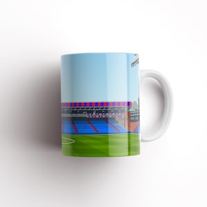 May include: White ceramic mug featuring a colourful illustration of a football stadium. The stadium has blue and red seats and a green pitch.