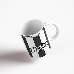May include: White ceramic mug with a black and white striped design. The design features a black and white soccer jersey with the word "sela" printed on it.