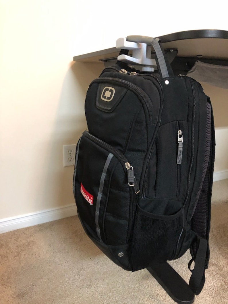 May include: A black backpack with a red "Makita" logo hanging from a white plastic hook attached to a desk.