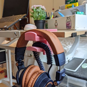 May include: A pair of brown and blue headphones with a coiled cord are hanging on a pink 3D printed headphone stand. The stand is attached to a wooden desk.
