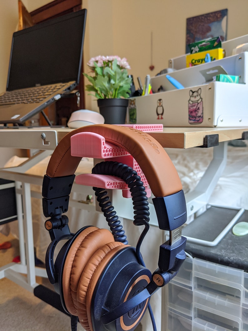 May include: A pair of brown and blue headphones with a coiled cord are hanging on a pink 3D printed headphone stand. The stand is attached to a wooden desk.