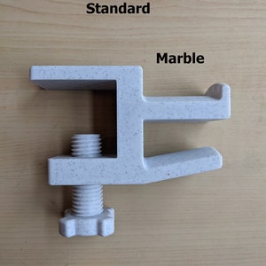 May include: A white plastic clamp with a speckled finish, labeled "Standard" and "Marble". The clamp has a screw with a hexagonal head.
