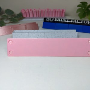 May include: A pink rectangular sign with two small white circles on each end. The sign is sitting on a white surface.