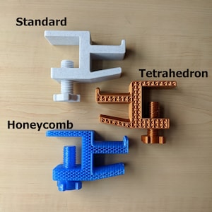 May include: Three 3D printed desk clamps in different colors and patterns. The white clamp is labeled "Standard", the orange clamp is labeled "Tetrahedron", and the blue clamp is labeled "Honeycomb".