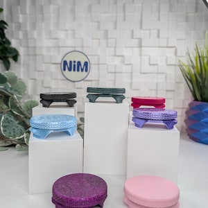 May include: A collection of circular, textured stands in various colors including black, dark green, light blue, purple, magenta, and pink. Each stand features a flat top and a base with three small legs. A circular logo with the text NiM is visible.