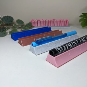 May include: Four 3D printed pen holders in different colors: blue, brown, white with black speckles, and pink. The holders are designed to hold pens and markers upright. The pink holder has the text "5D PRINT FACTORY" printed on it.