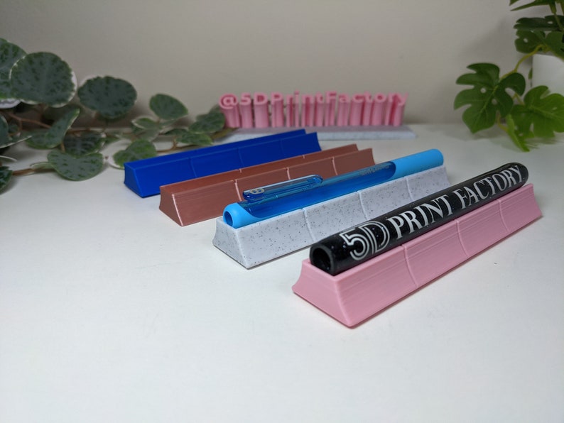 May include: Four 3D printed pen holders in different colors: blue, brown, white with black speckles, and pink. The holders are designed to hold pens and markers upright. The pink holder has the text "5D PRINT FACTORY" printed on it.