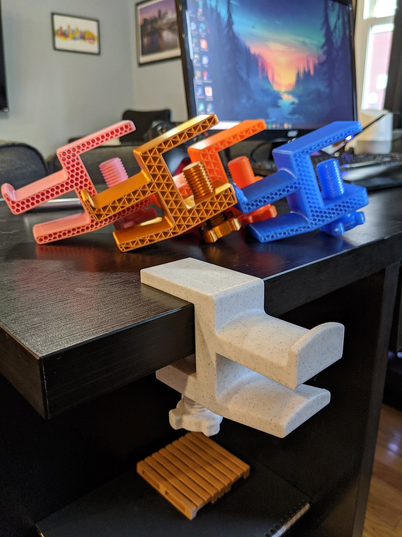 May include: Four 3D printed desk clamps in different colors: pink, orange, blue, and white. The clamps are designed to hold items like phones, tablets, or other devices.